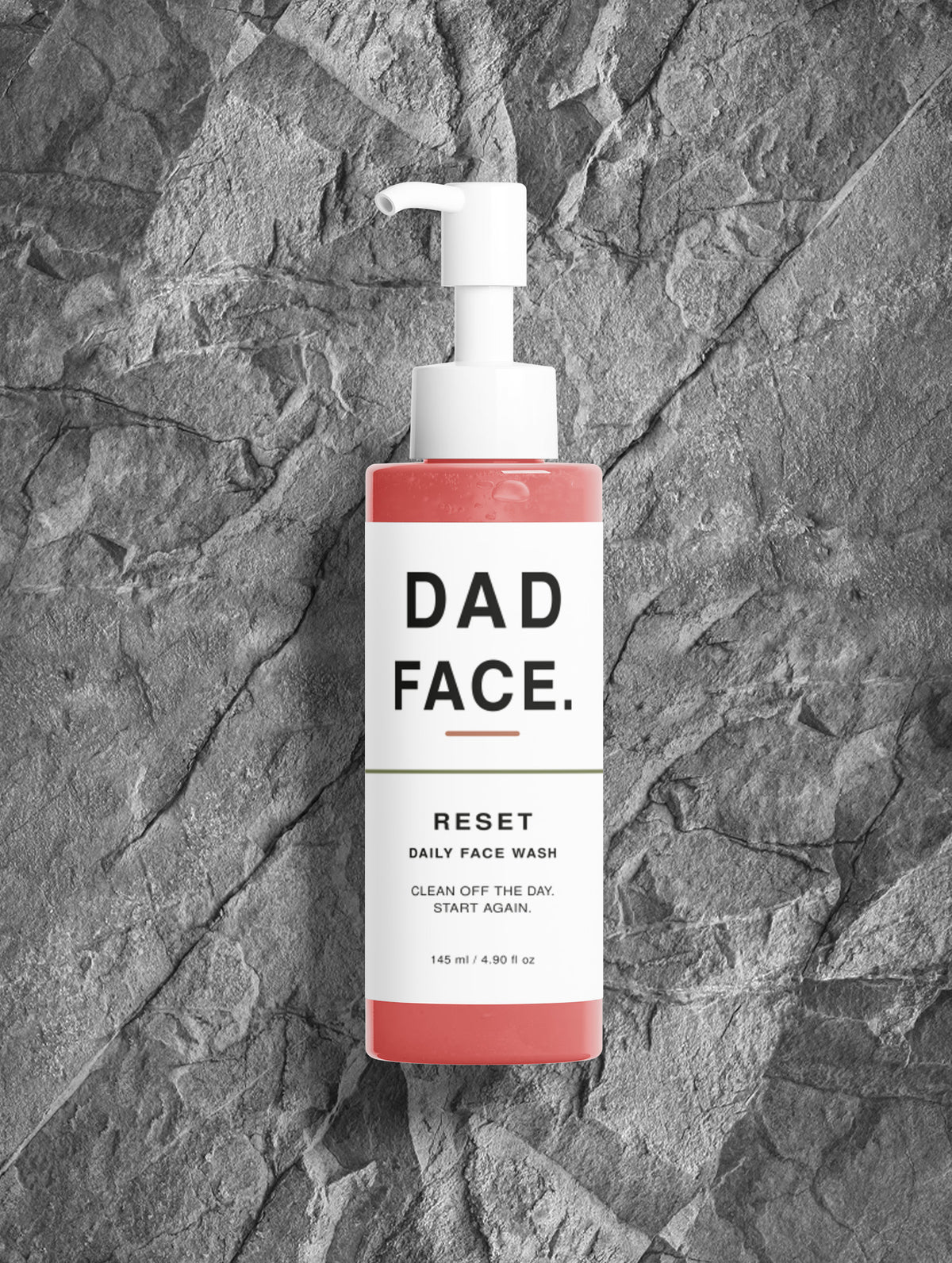 A bottle of Dad Face Reset Daily Face Wash with a white pump and a white label, featuring coral pink liquid at the top and bottom, set against a dark gray textured stone background.