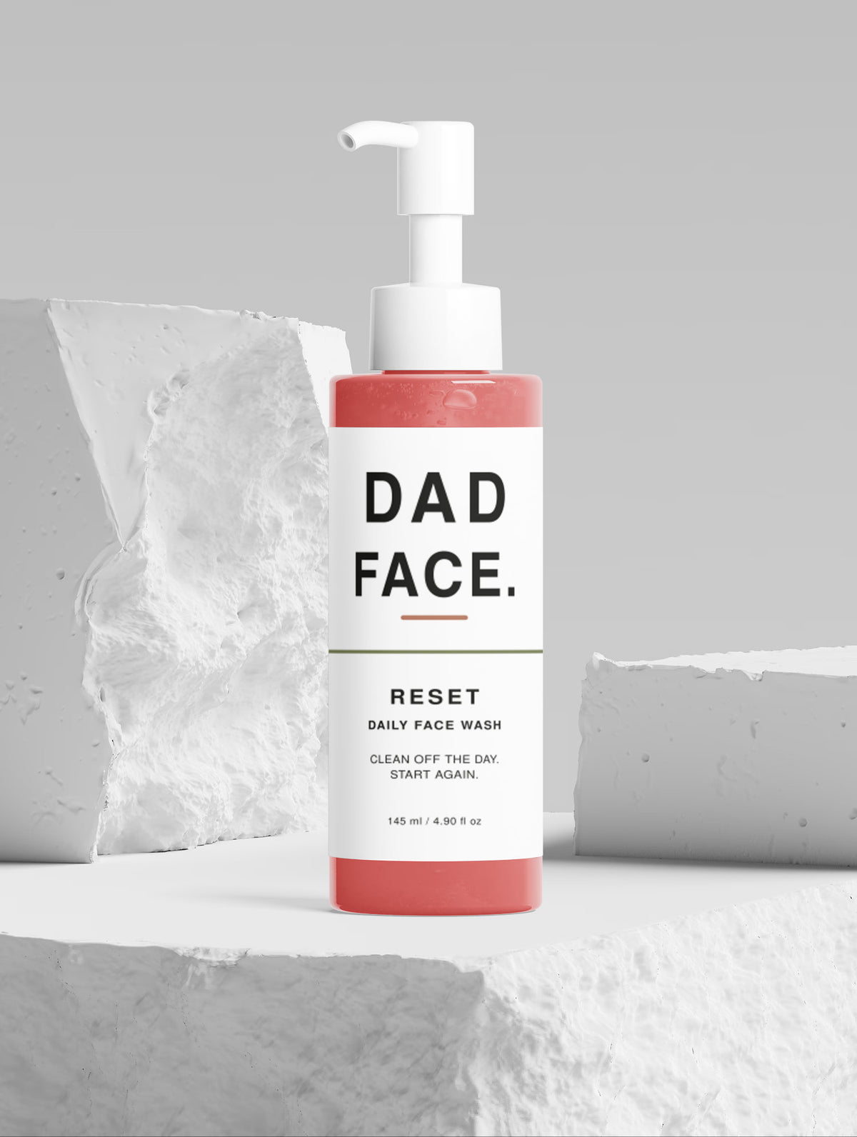 A bottle of Dad Face Reset Daily Face Wash stands on a white pedestal against a gray background with textured white stone props. The bottle has a coral-red base, a white label with black text, and a white pump dispenser.