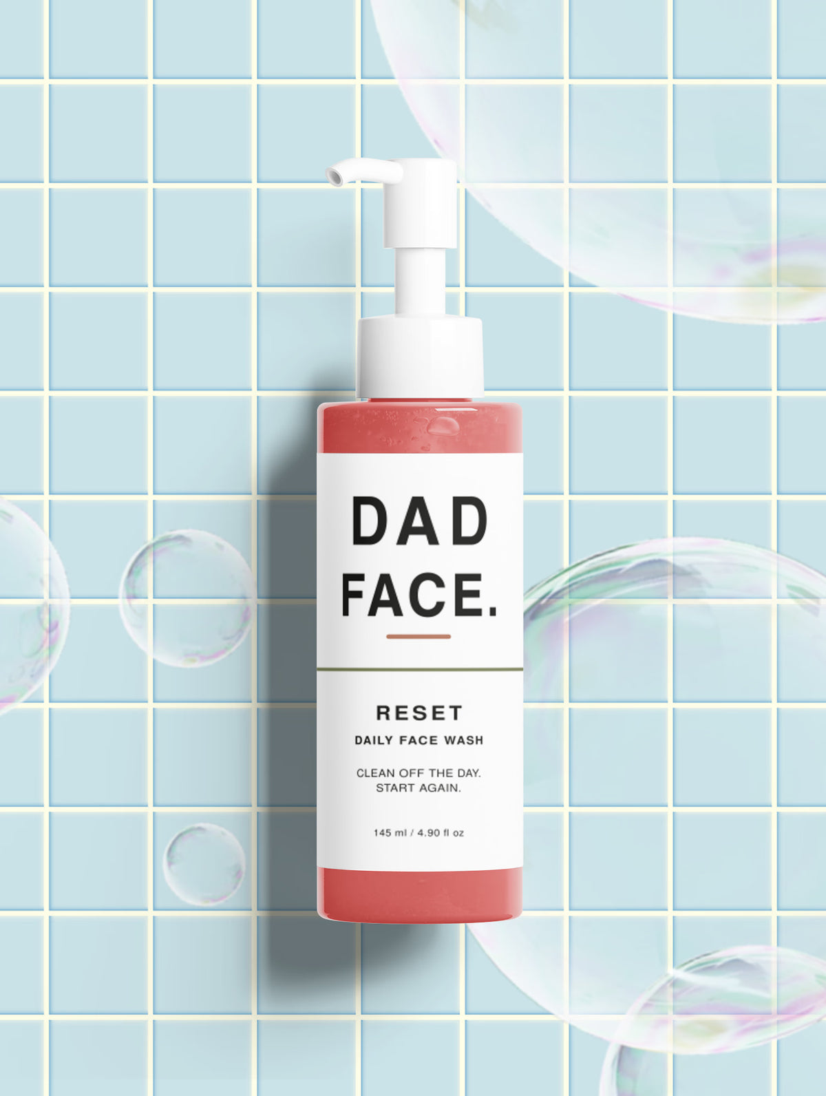 A bottle of Dad Face Reset Daily Face Wash stands against a light blue tiled wall with floating soap bubbles. The bottle has a coral pink lower half and a white upper half with black text.
